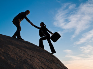 A silhouette of two business people helping each other up a cliff.